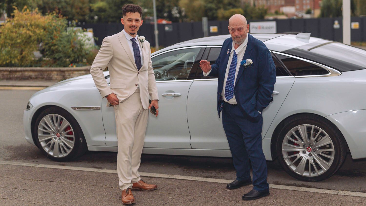 Two men in suits posing beside a white Jaguar luxury car outdoors on a sunny day.