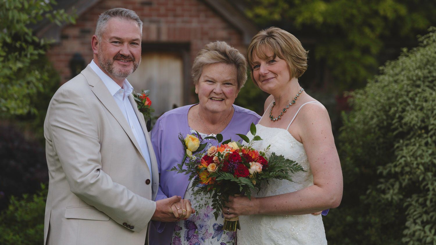 A bride and groom pose with an older woman holding a colorful bouquet outdoors at a wedding celebration.