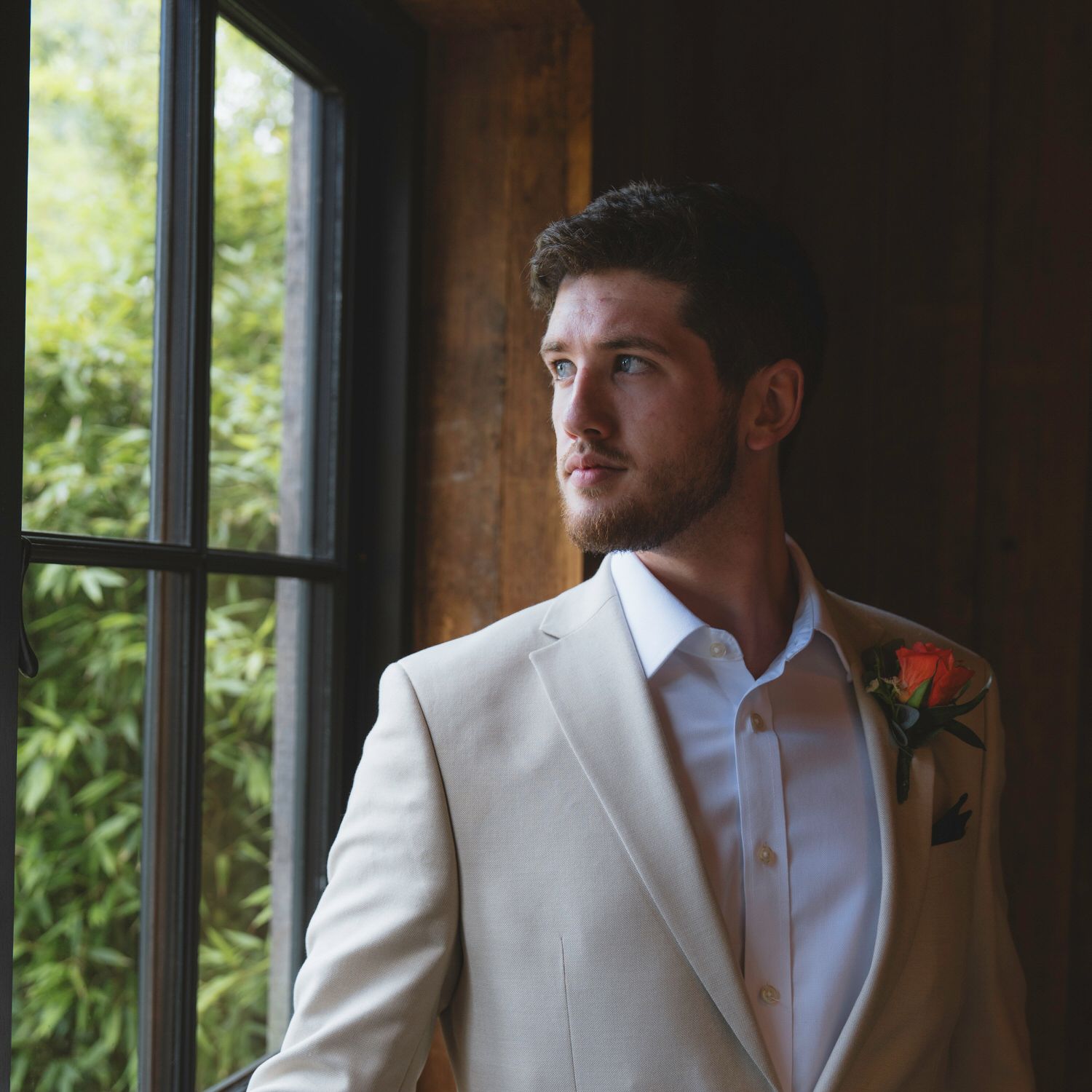 A young groom in a cream suit with a floral boutonniere gazes thoughtfully out a window in soft natural light.