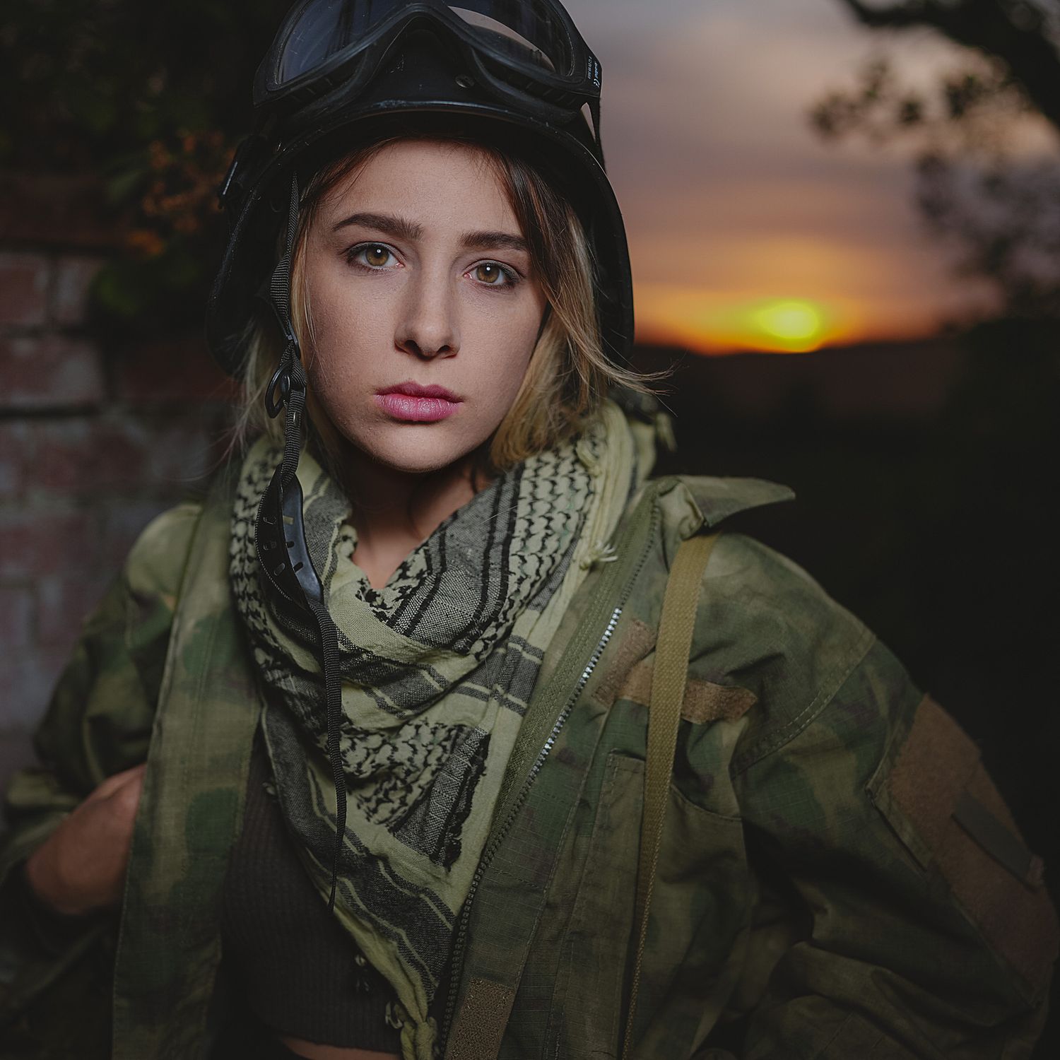 Woman in camouflage jacket and keffiyeh scarf wearing black helmet at golden sunset.