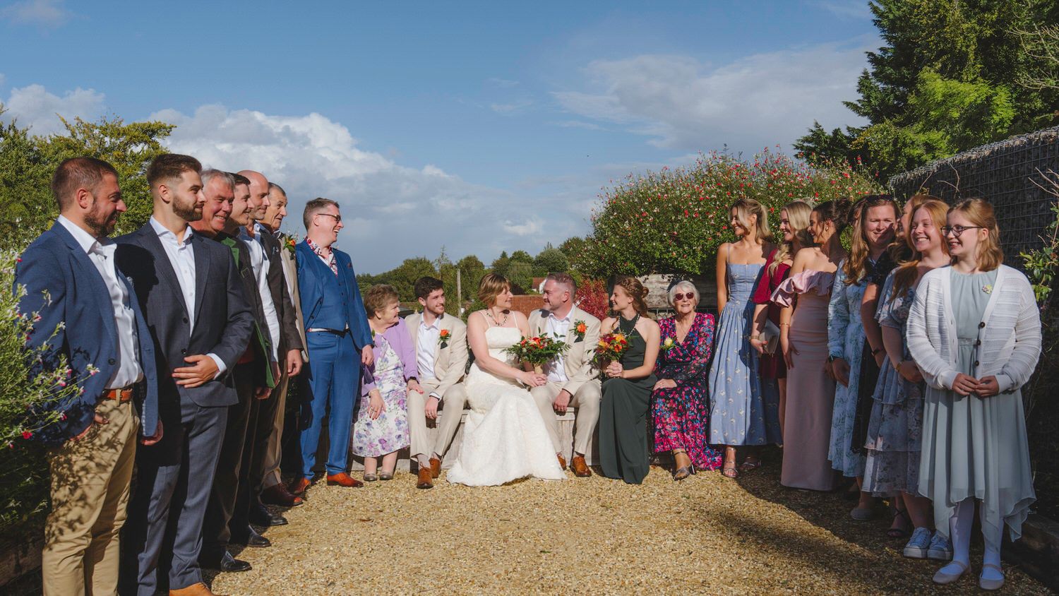 Wedding party group photo outdoors, bride and groom surrounded by guests in formal attire on a sunny day.