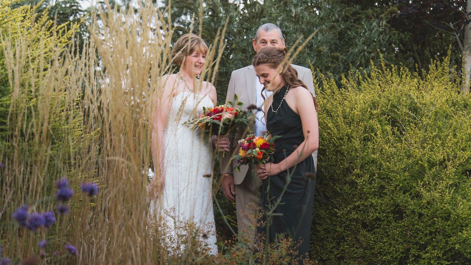 Bride and groom with wedding guest admiring bouquets among tall grasses and wildflowers in a garden ceremony.