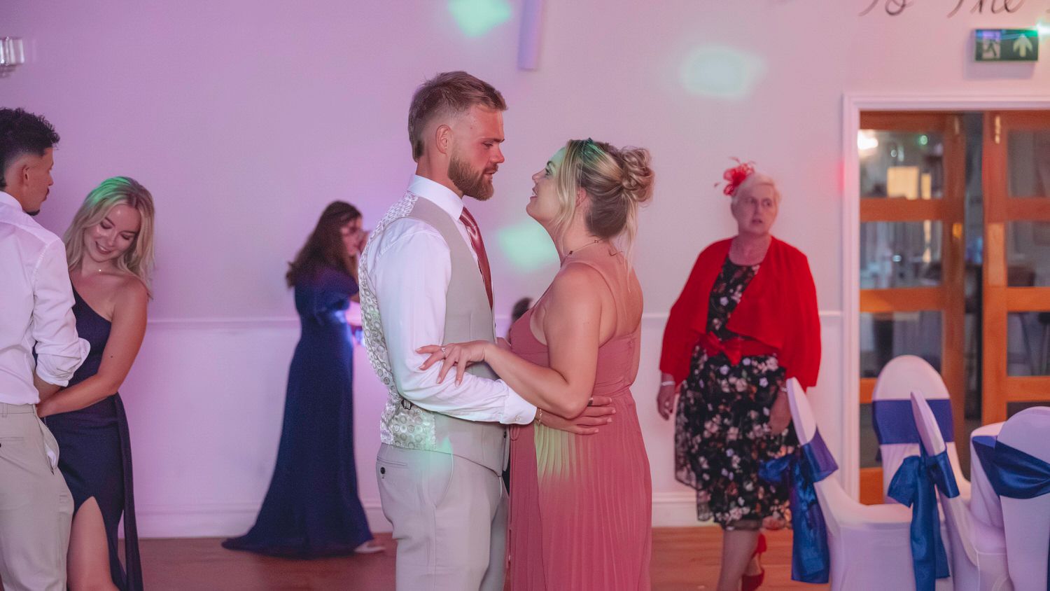 Couple sharing a romantic dance at a wedding reception, surrounded by guests in formal attire in an elegant venue.