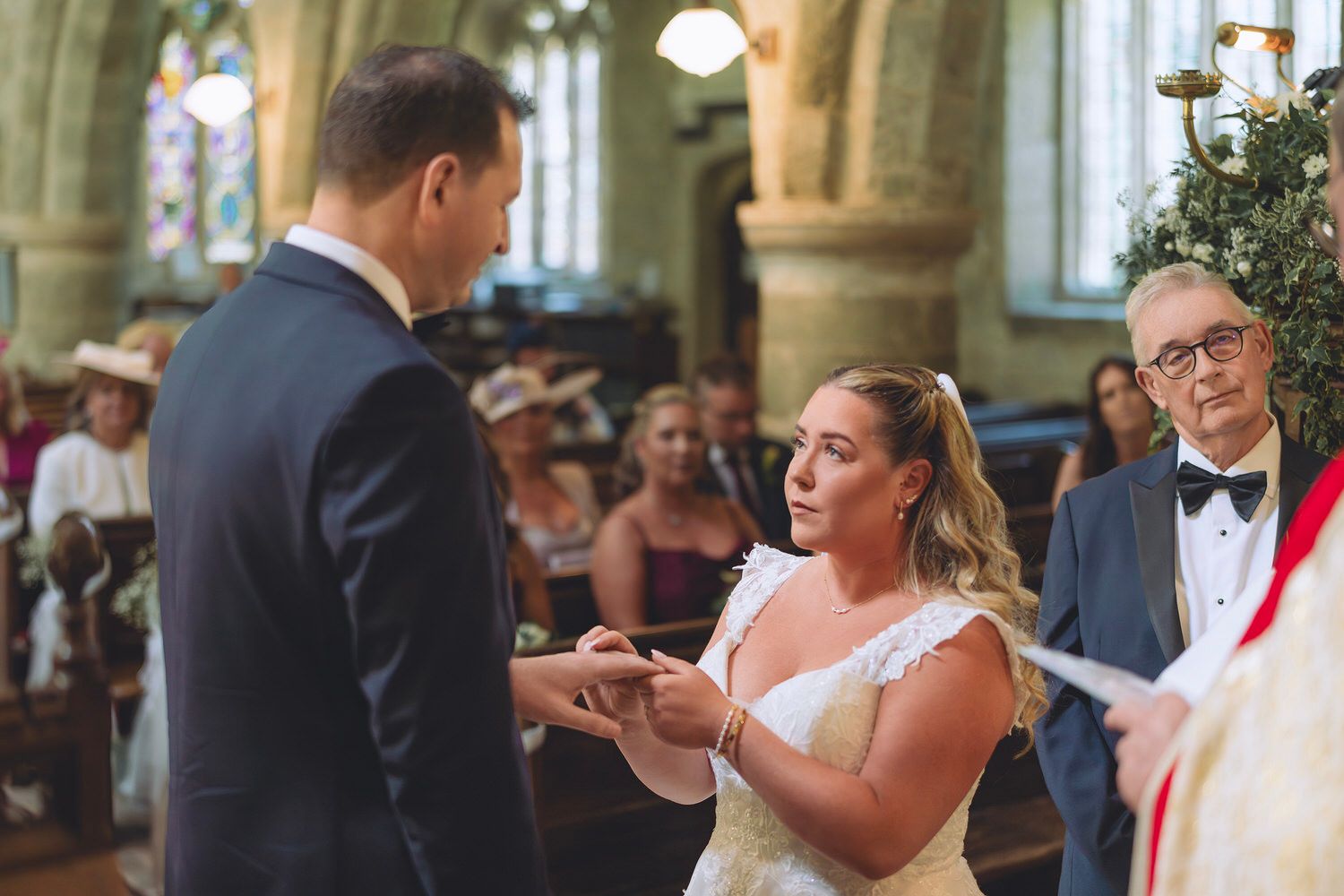 Bride placing wedding ring on groom's finger during church ceremony with guests seated in pews.