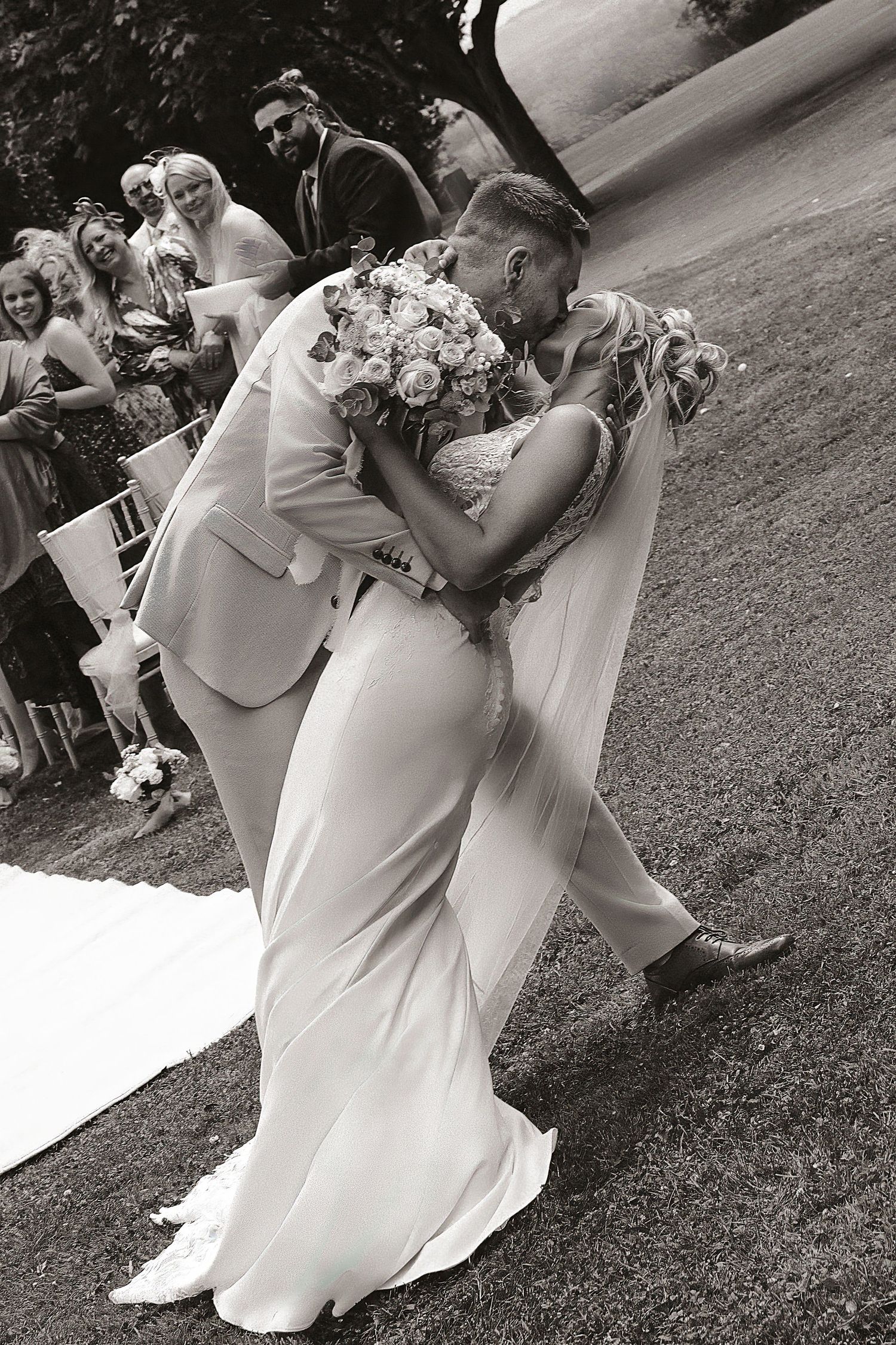 Black and white photo of bride and groom sharing first kiss at outdoor wedding ceremony with guests watching.