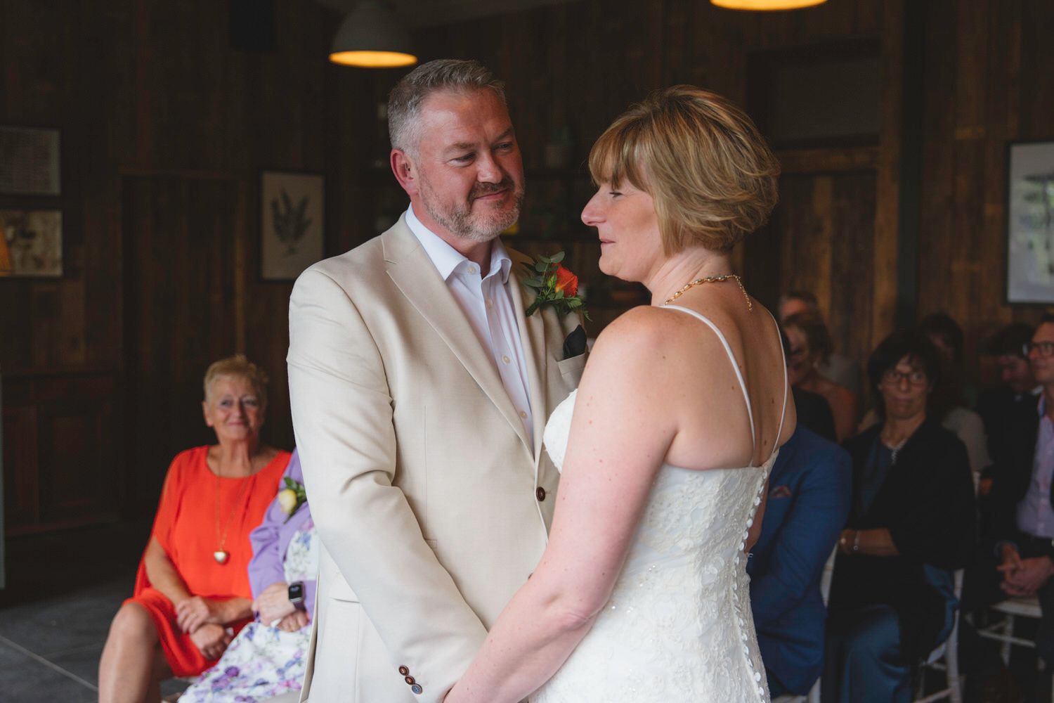 Bride and groom exchange vows at indoor wedding ceremony, guests seated in background.