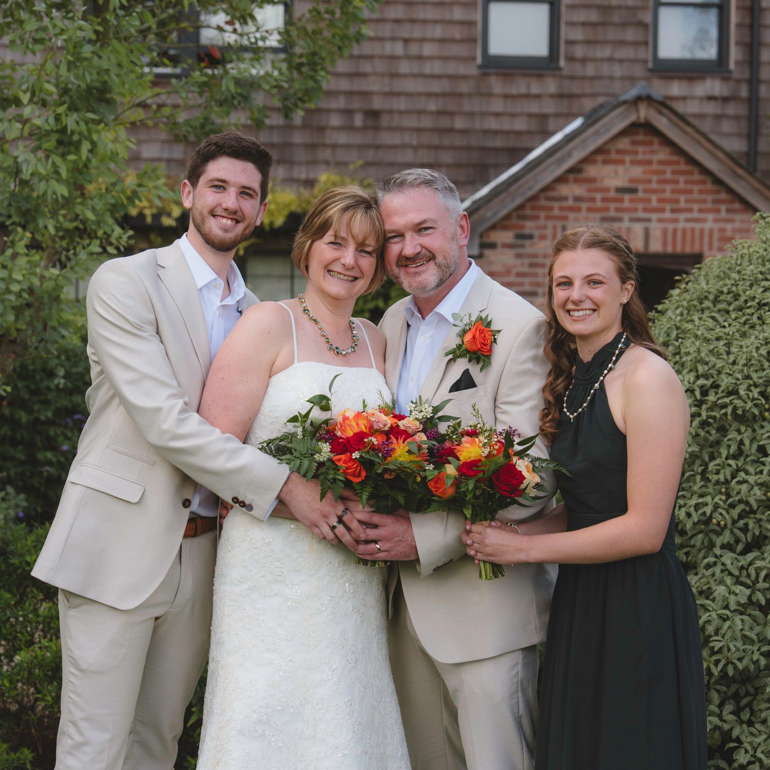 Happy wedding couple with family posing outdoors, bride holding colorful bouquet, guests in formal attire near brick building.
