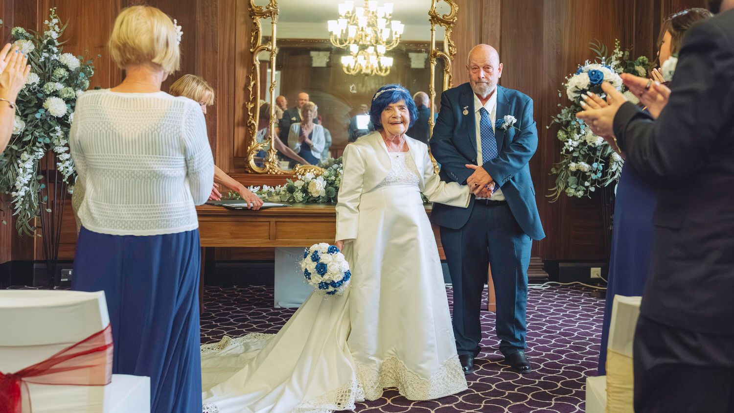 Joyful wedding couple walks down the aisle, bride in white gown, groom in blue suit, guests applauding in elegant venue.