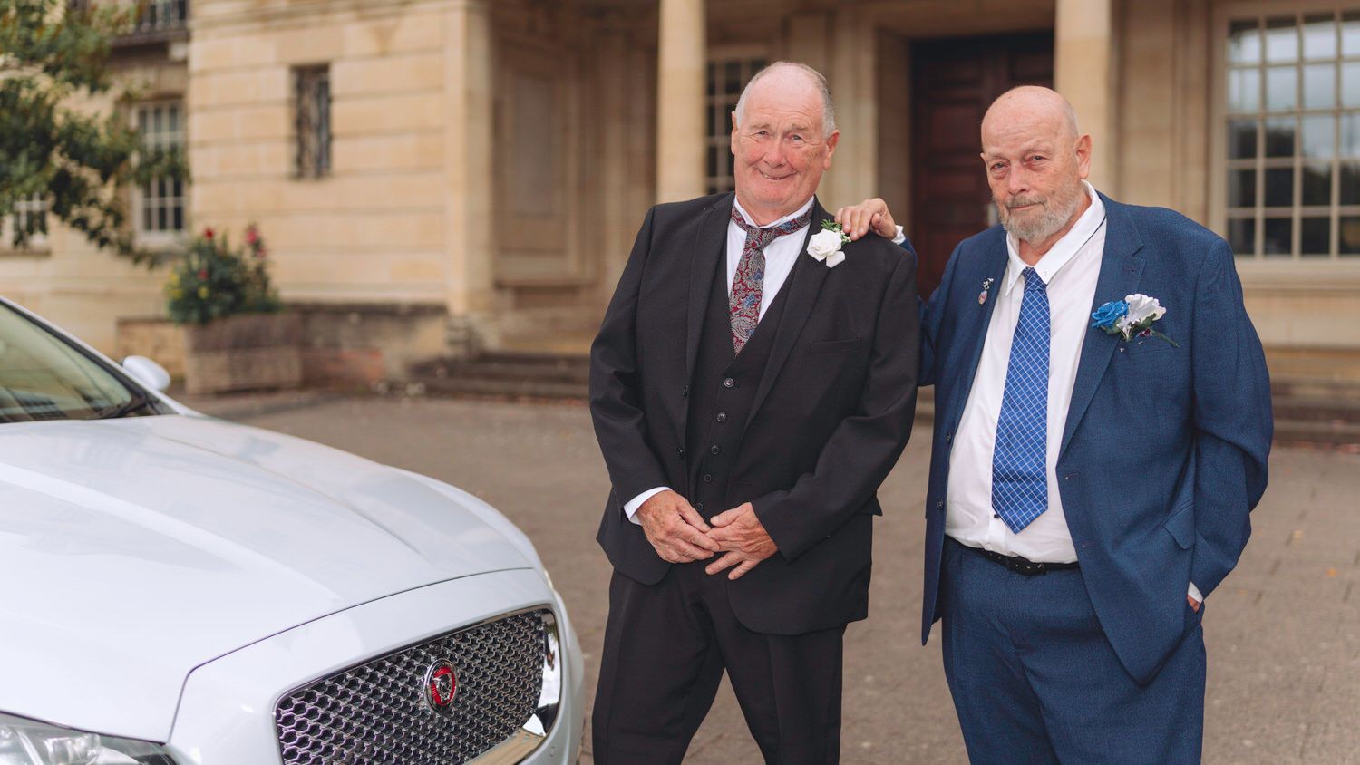 Two elderly men in formal suits pose beside a white Jaguar car outside an elegant building at a wedding.