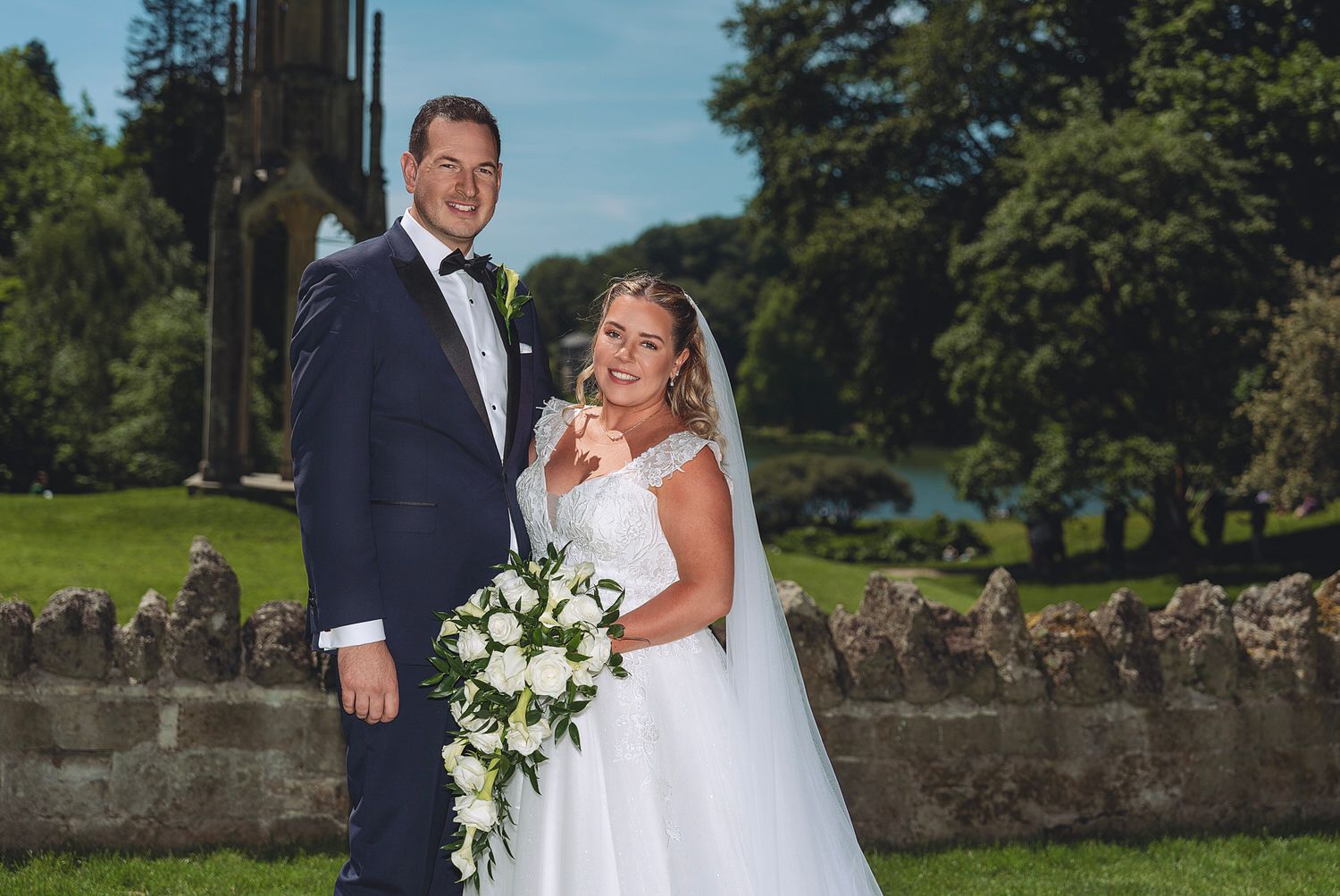Bride and groom pose outdoors in lush garden setting, bride holding white floral bouquet in elegant wedding gown.