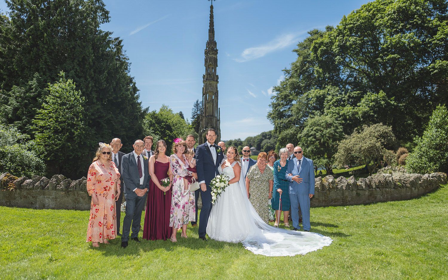 Wedding group photo with bride and groom surrounded by family in a sunny garden with a stone monument backdrop.