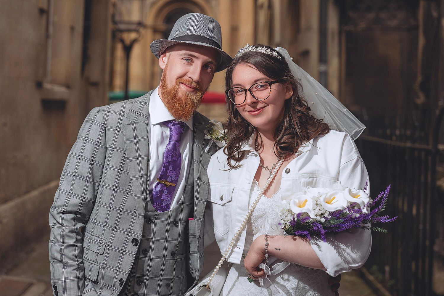 Smiling bride and groom posing together on their wedding day, bride holding purple and white bouquet.