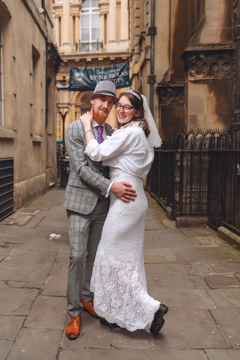 A bride and groom embrace on a cobblestone alley, she in lace gown, he in plaid suit with red beard and hat.