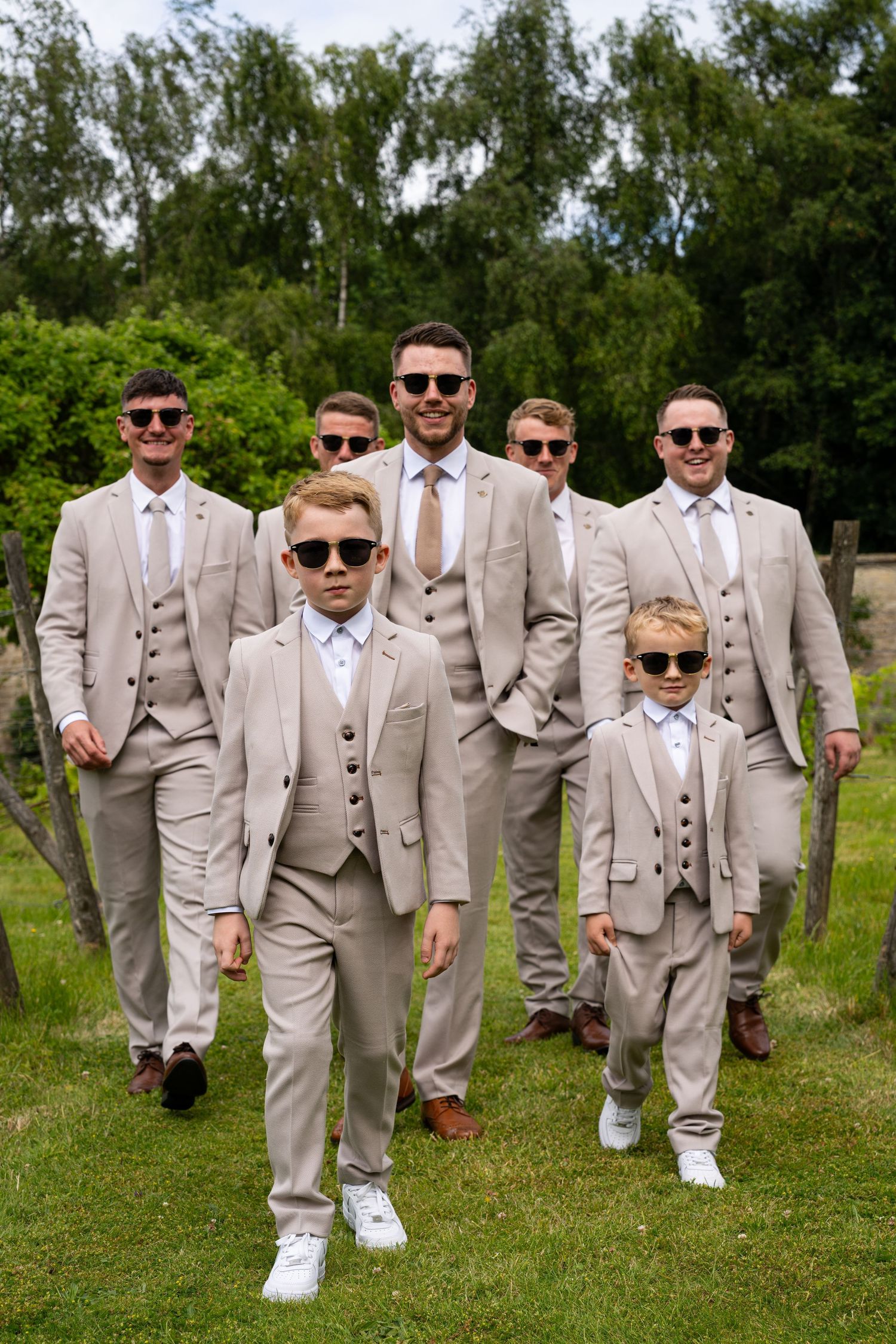 Groomsmen and ring bearers in matching beige suits and sunglasses walking outdoors at a wedding.