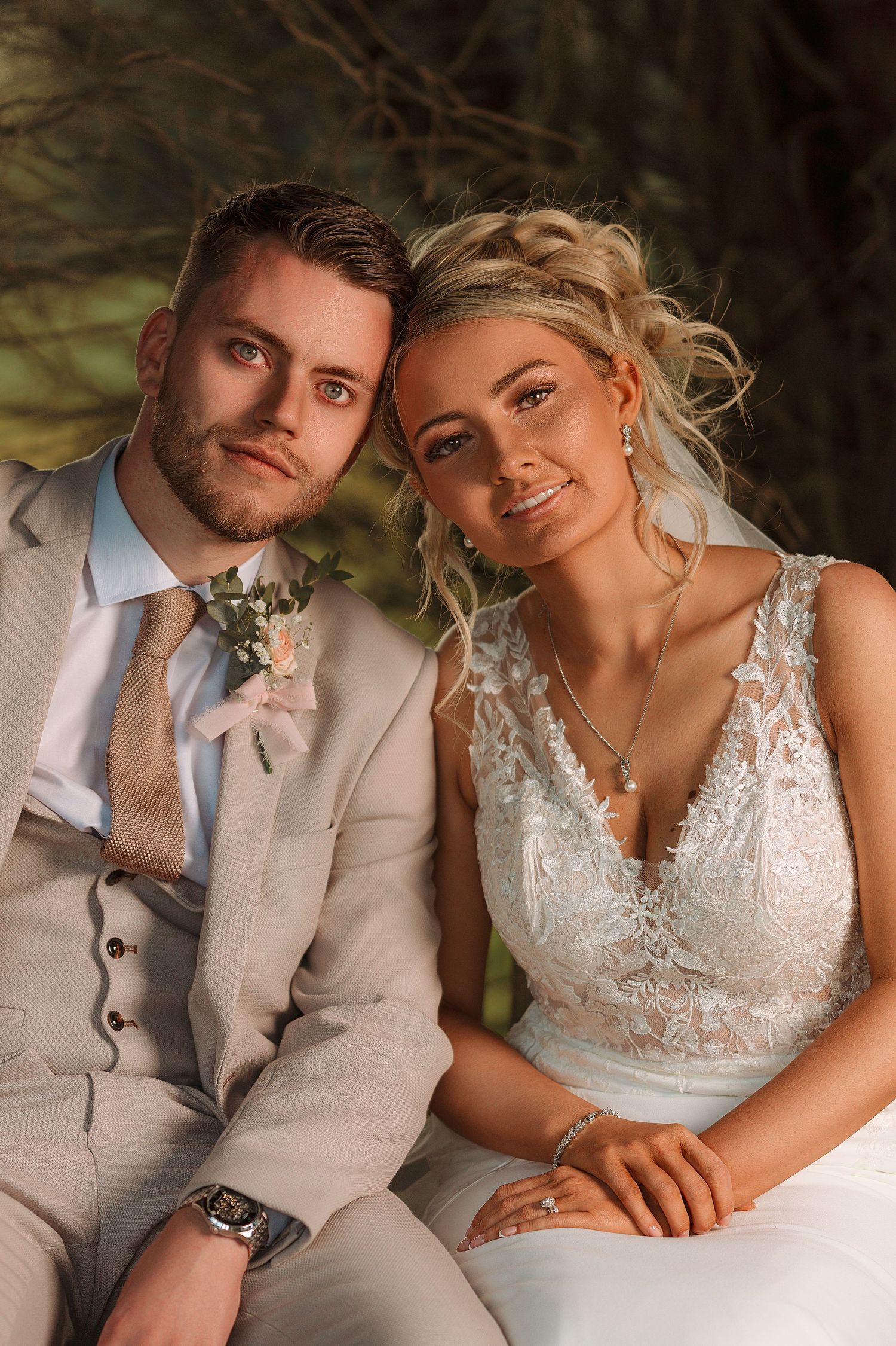 A bride and groom pose together outdoors, she in white lace gown, he in beige suit with floral boutonniere.