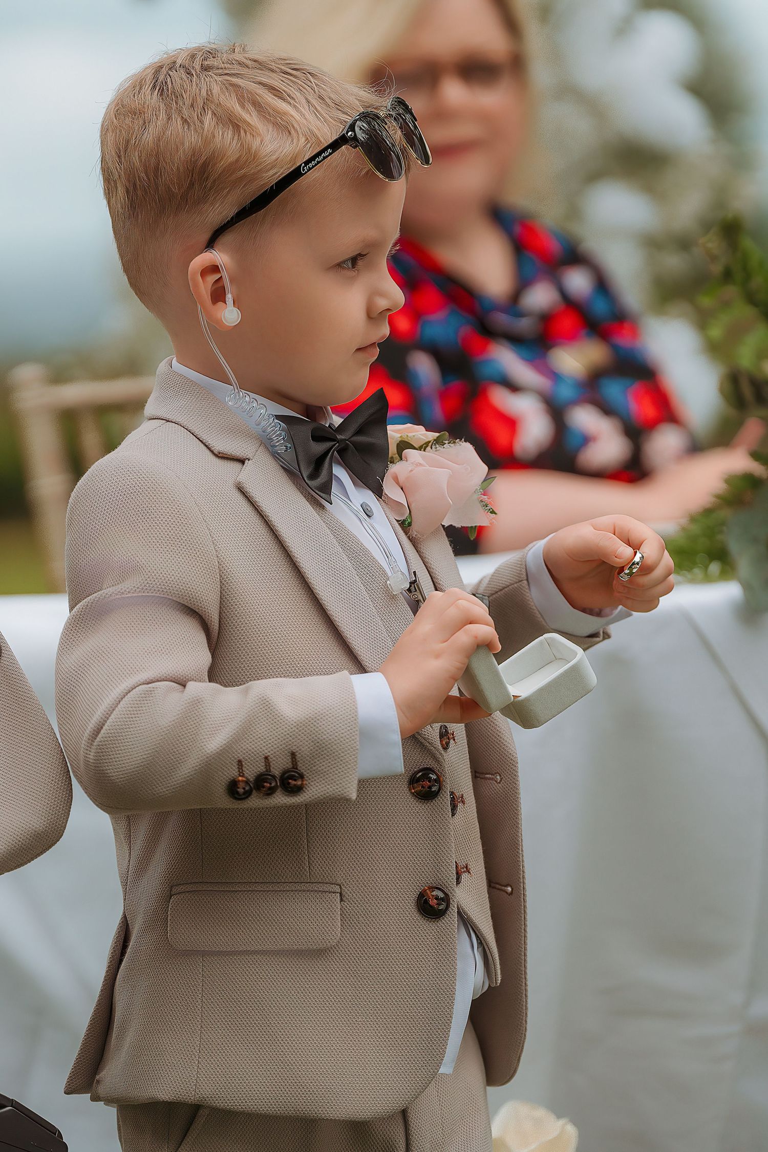 Young ring bearer in tan suit, bow tie, and sunglasses holds a ring pillow at an outdoor wedding ceremony.