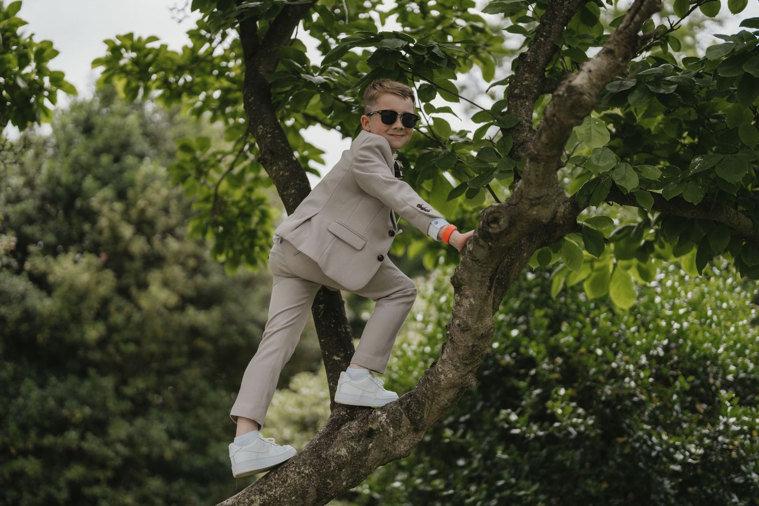 A young boy in a beige suit and sunglasses climbs a tree branch in a lush green garden.