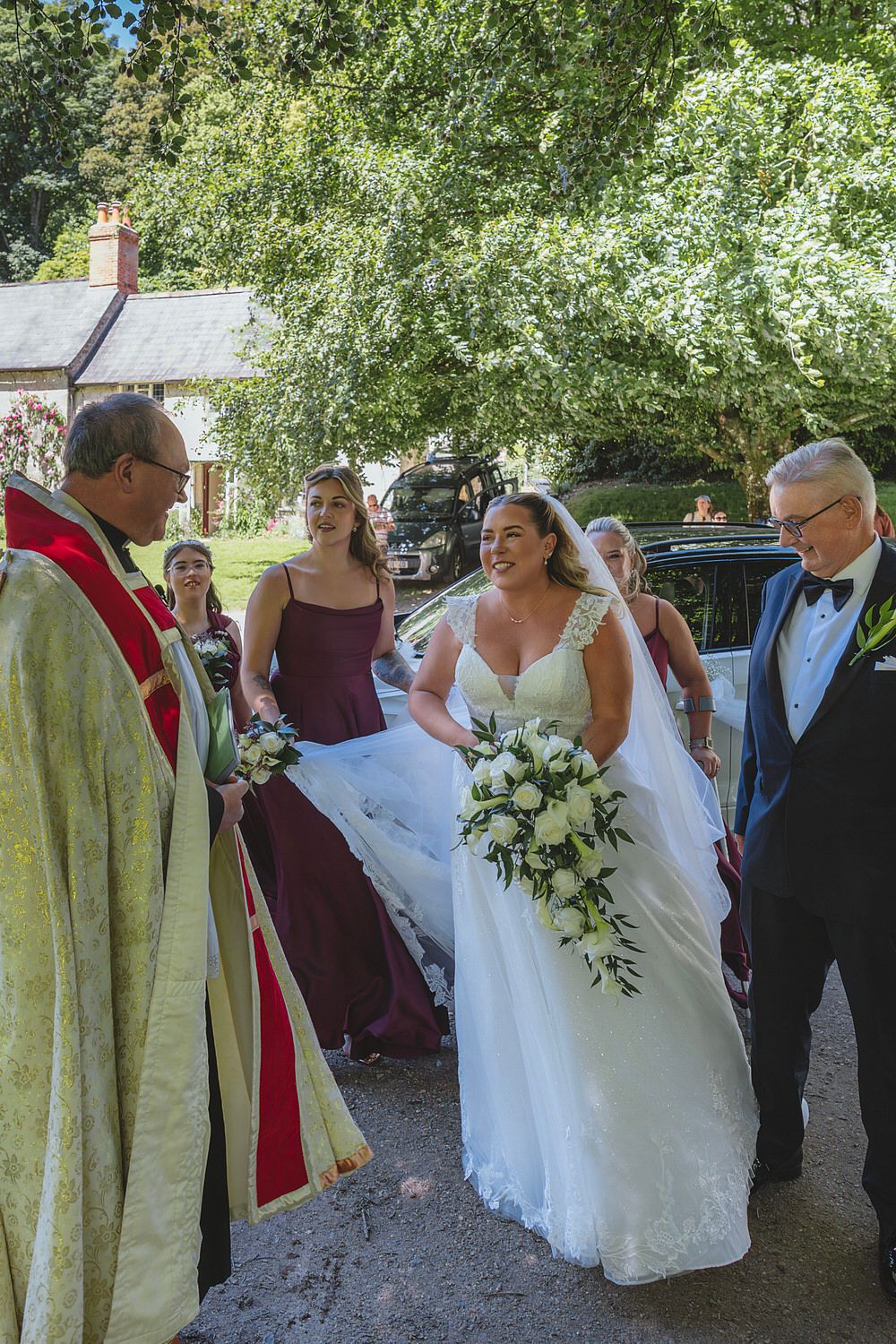 Bride in white gown with bouquet greeted by officiant, flanked by bridesmaids in burgundy and elderly gentleman in tuxedo.