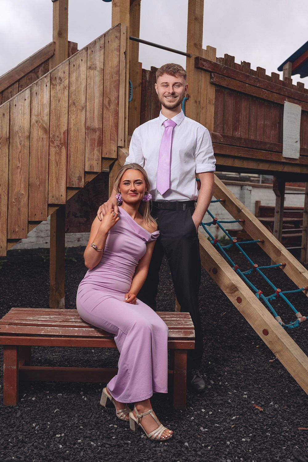 A young couple dressed in matching lavender outfits poses near a wooden playground structure for a formal occasion.