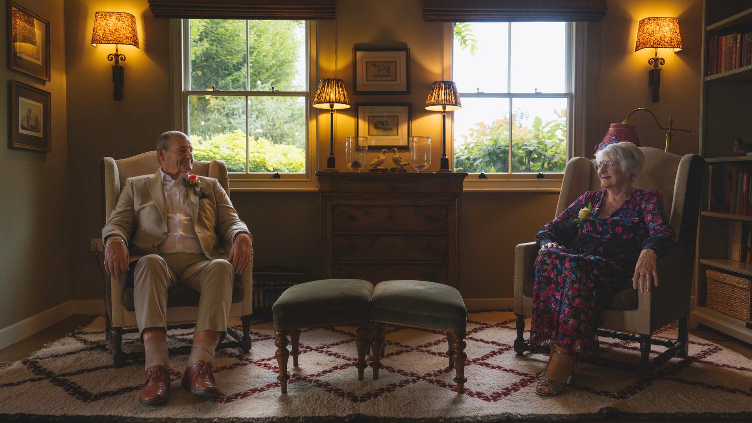 Elderly couple sitting in armchairs in a warmly lit living room with vintage decor and a large window.