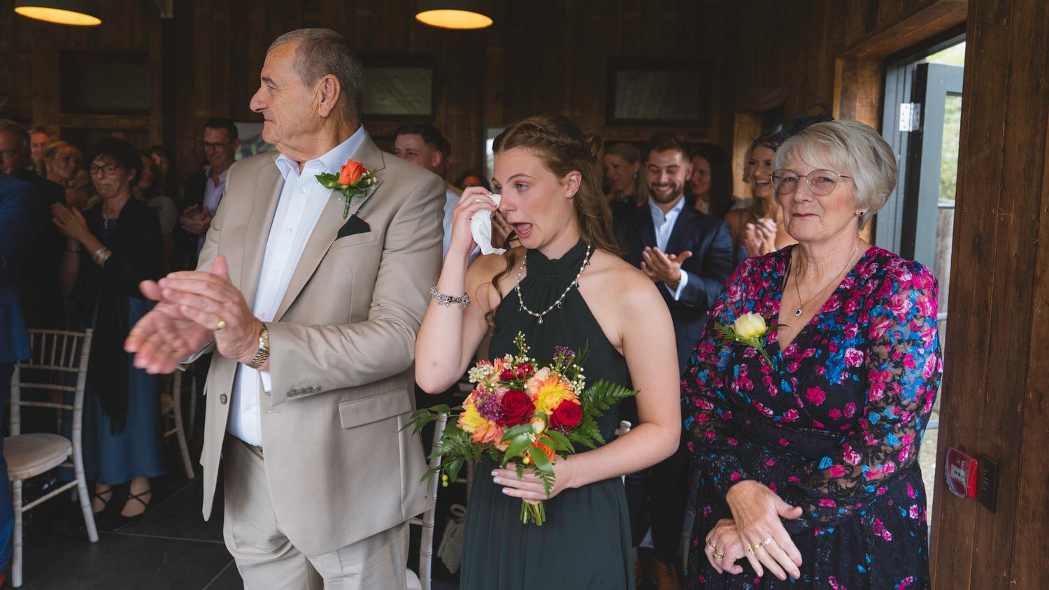 Emotional bridesmaid wiping tears holding bouquet flanked by family at rustic wedding ceremony.