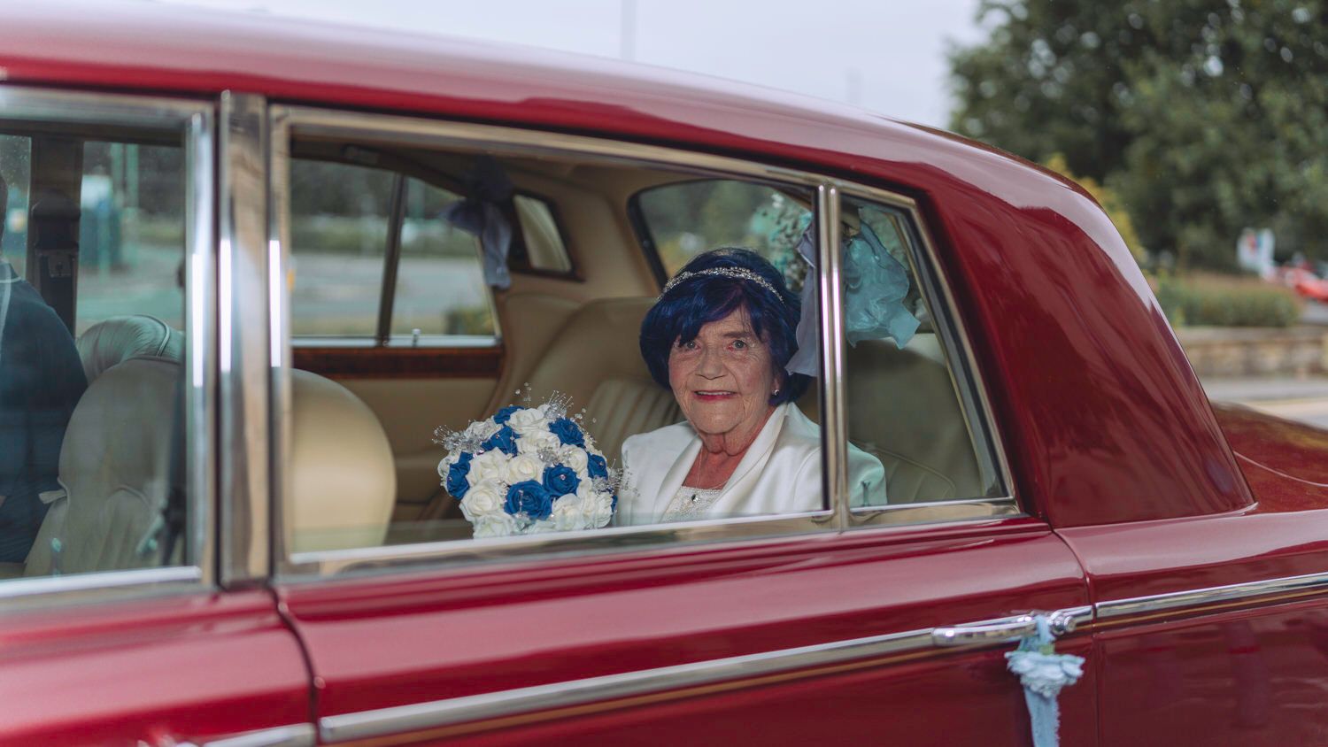 Elderly bride sitting in a classic red car holding a blue and white bouquet, looking out the window.