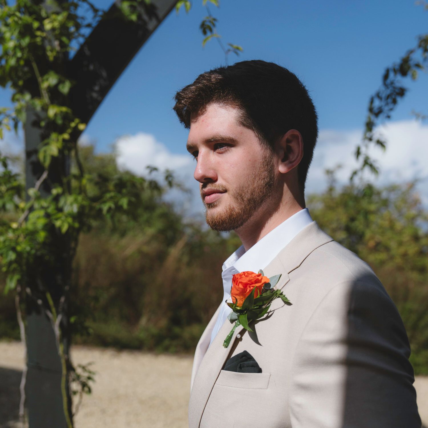 A groom in a beige suit with an orange boutonniere stands outdoors under a floral arch on a sunny day.