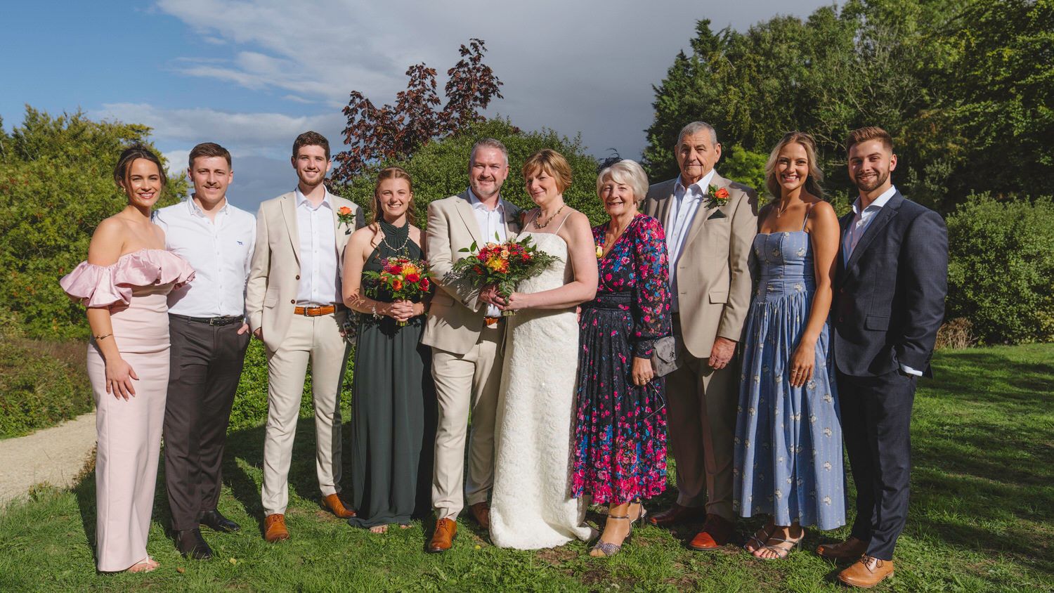Wedding party group photo outdoors, bride in white gown with bouquet, groom in tan suit, family in formal attire.