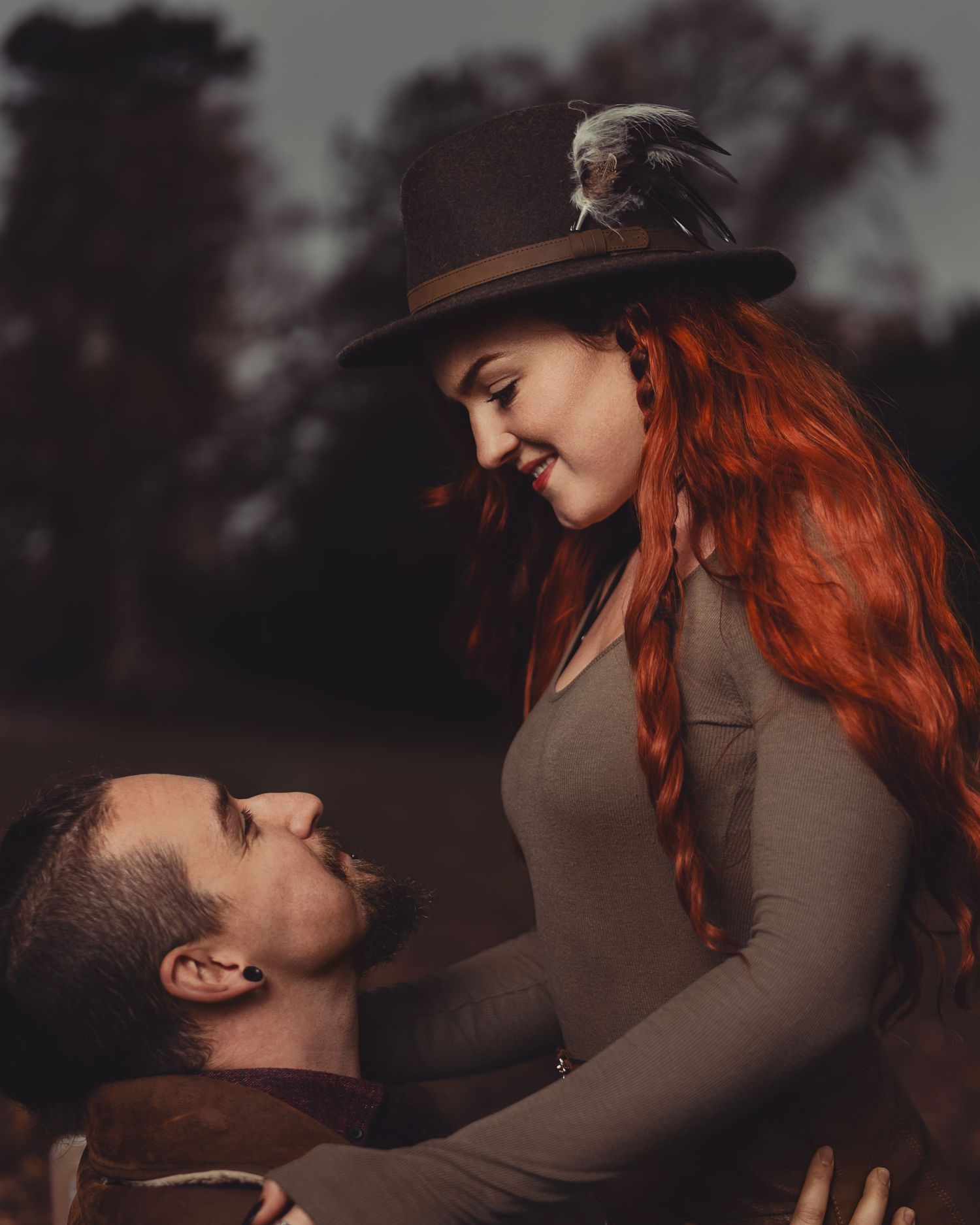 Smiling red-haired woman in a feathered brimmed hat gazes down at a bearded man looking up at her in a moody autumn outdoor portrait.