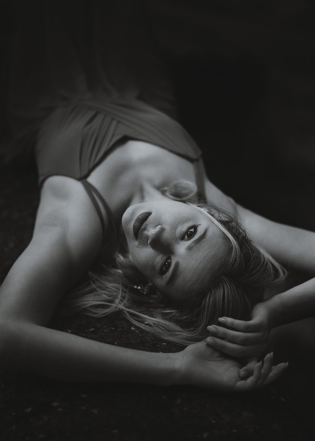 Black and white portrait of a blonde woman lying on the ground in a dress, gazing upward dramatically.
