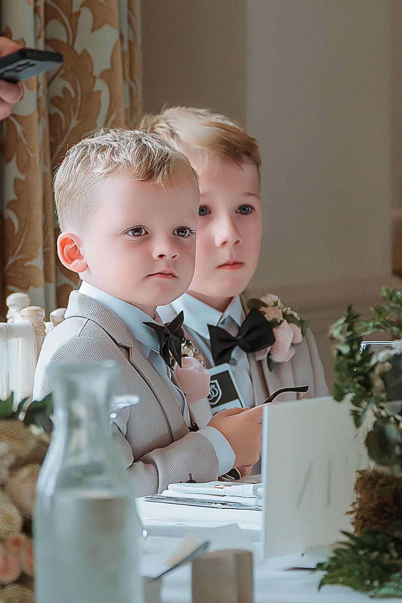 Two young boys in matching tan suits with bow ties seated at a wedding reception table.