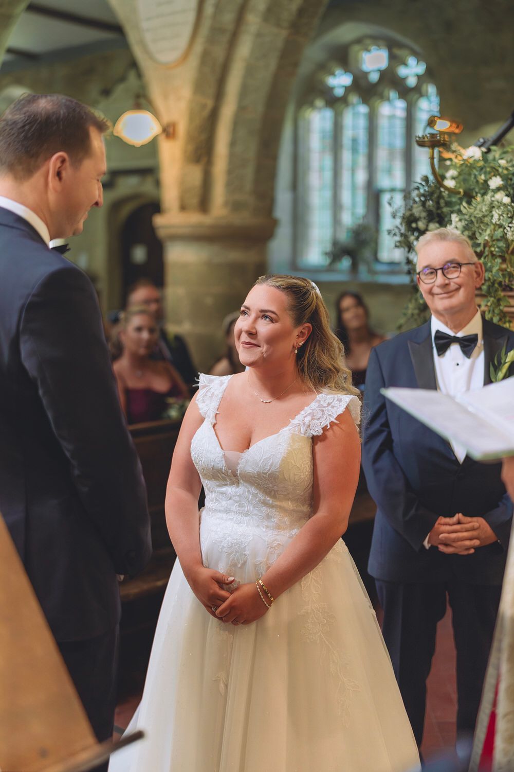Bride smiling joyfully at groom during wedding ceremony in a historic church, wearing a lace A-line gown.