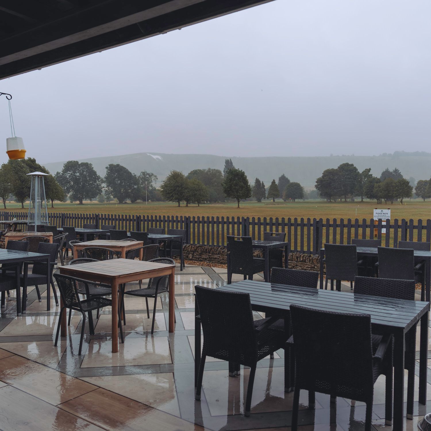 Wet outdoor patio with dark wicker chairs and tables overlooking a misty green golf course on a rainy day.