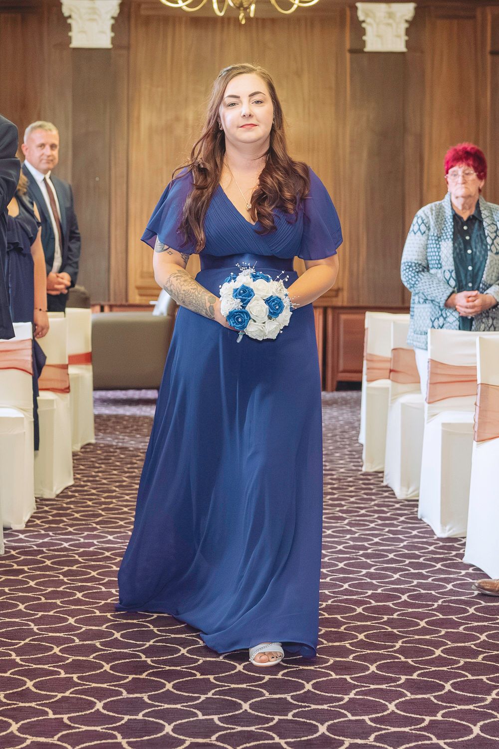 Bridesmaid in navy blue gown walks down aisle holding white and blue bouquet at elegant wedding ceremony.