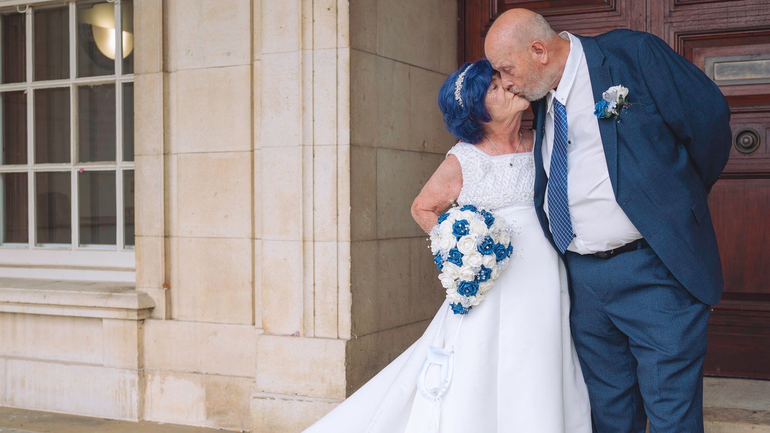 Couple sharing a kiss on their wedding day, bride in white gown with blue bouquet, groom in navy suit.