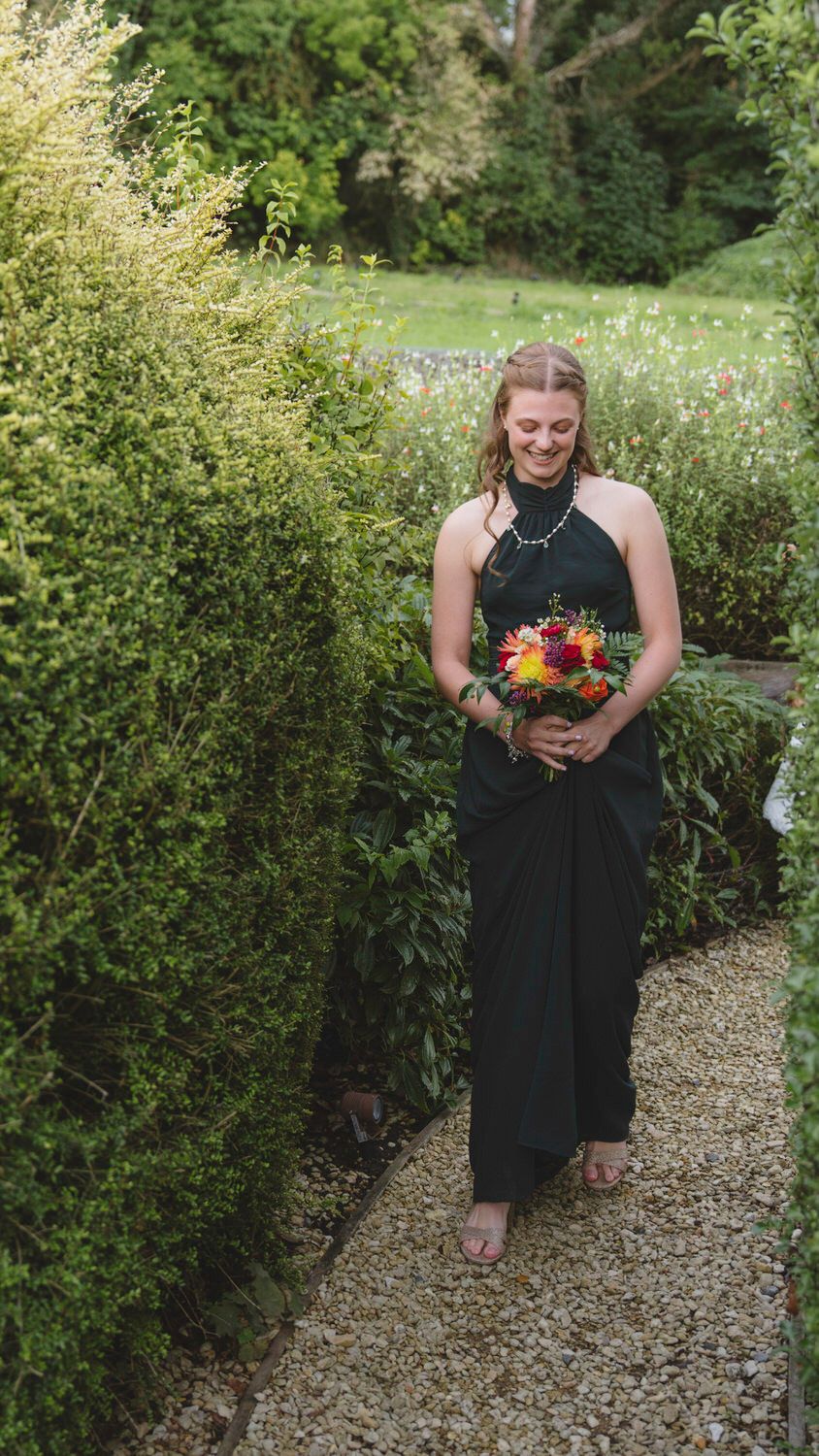 Bridesmaid in black halter gown holding colorful bouquet walks garden path lined with lush green hedges.
