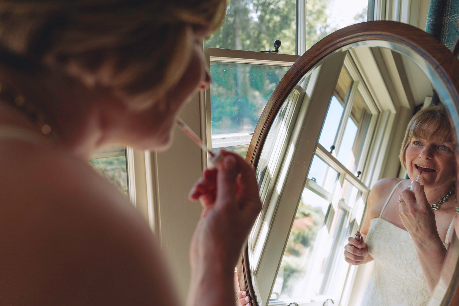 Bride applying makeup while smiling at her reflection in a vintage round mirror near a sunlit window on her wedding day.