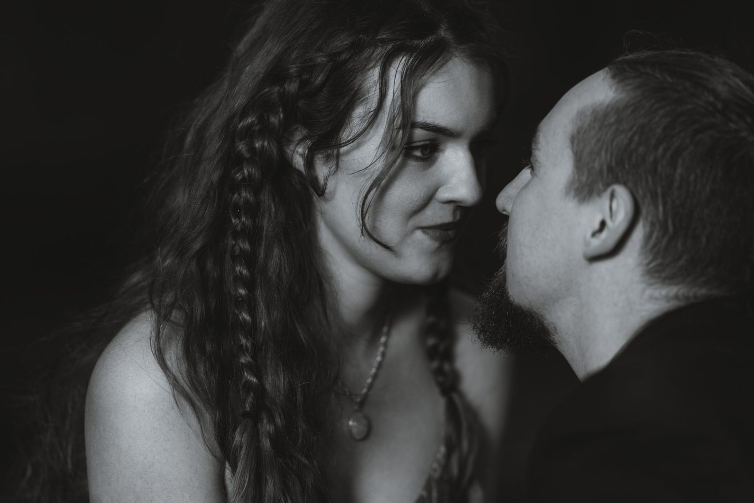 Black and white portrait of a woman with long braided hair and a pendant necklace leaning close to a man in a dark shirt.
