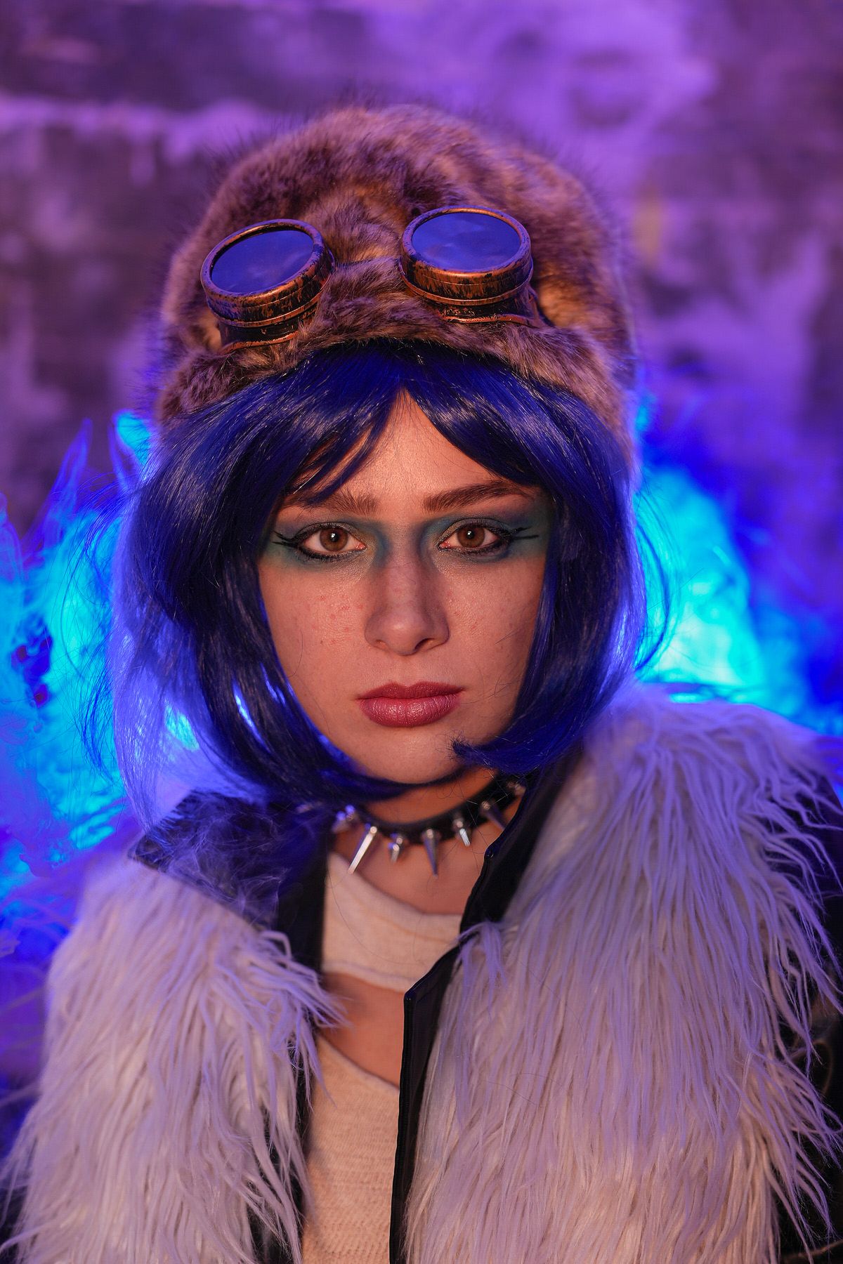 Woman with blue bob wig wearing fur hat with steampunk goggles and white fluffy coat in purple light.