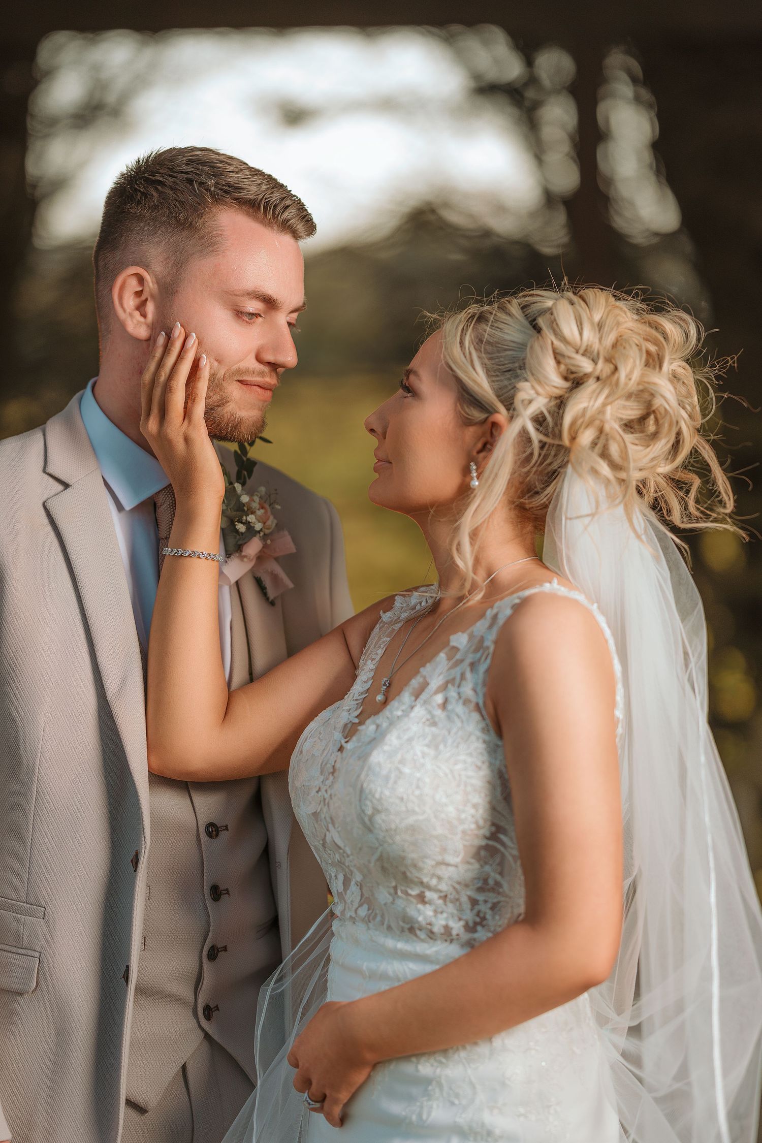 Bride tenderly touches grooms face as they share romantic moment in soft golden light outdoors.