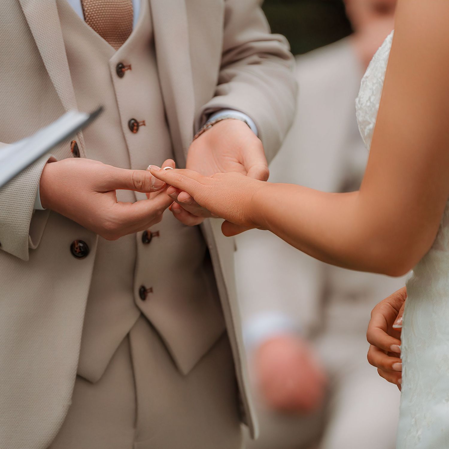 Groom placing a wedding ring on bride's finger during an outdoor ceremony, both dressed in elegant wedding attire.
