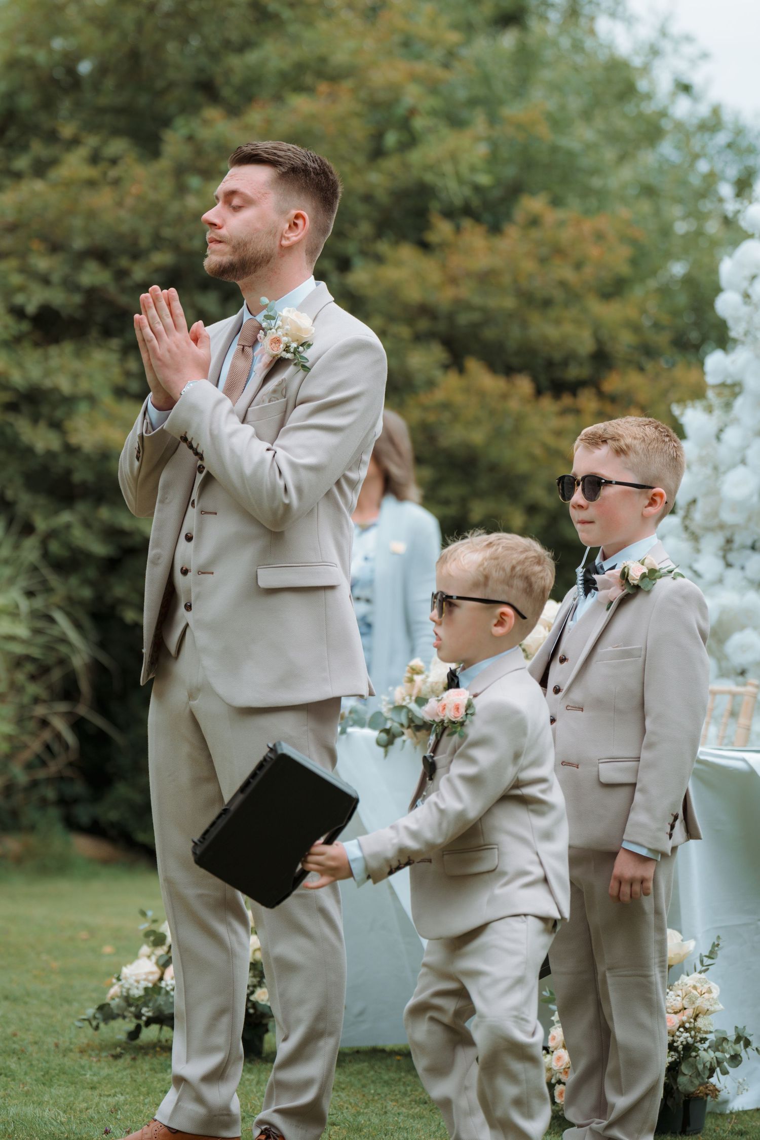 Groom in beige suit prays with hands together as two young ring bearers in matching suits and sunglasses stand beside him outdoors.