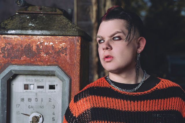 Person in orange striped sweater with edgy makeup and chain necklace posing beside a rusty vintage meter outdoors.