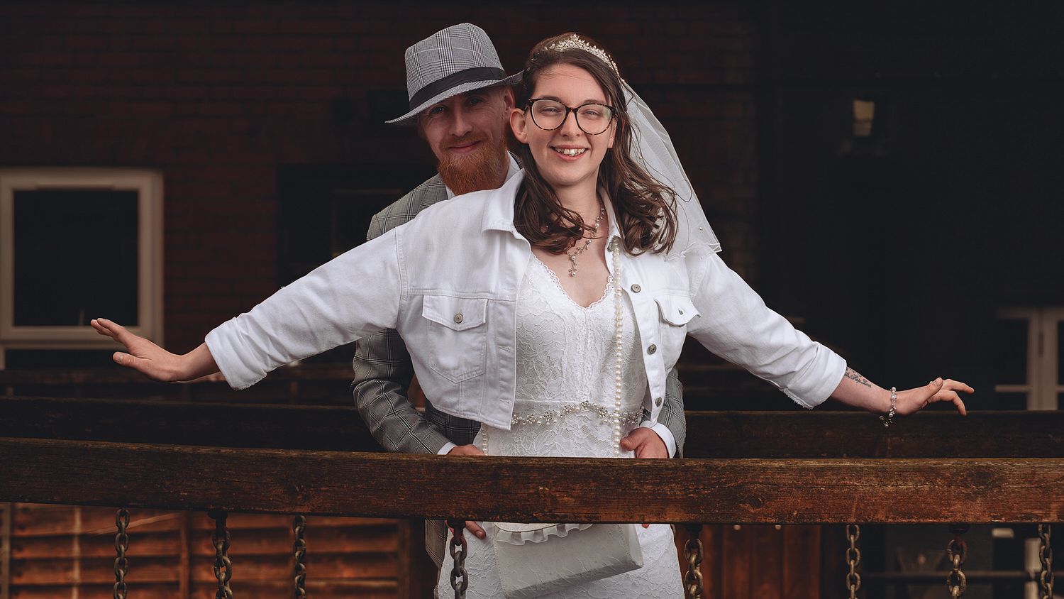 Couple in white wedding attire smiling on a wooden balcony, bride wearing veil and glasses.