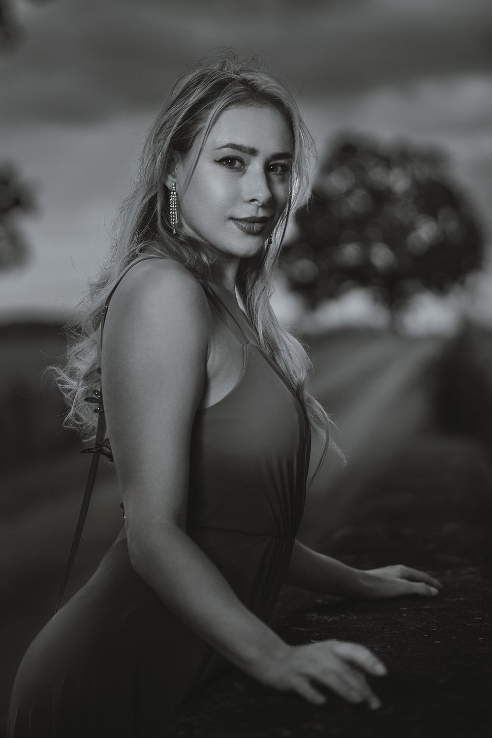 Black and white portrait of a young blonde woman in a sleeveless dress, posing outdoors at dusk.