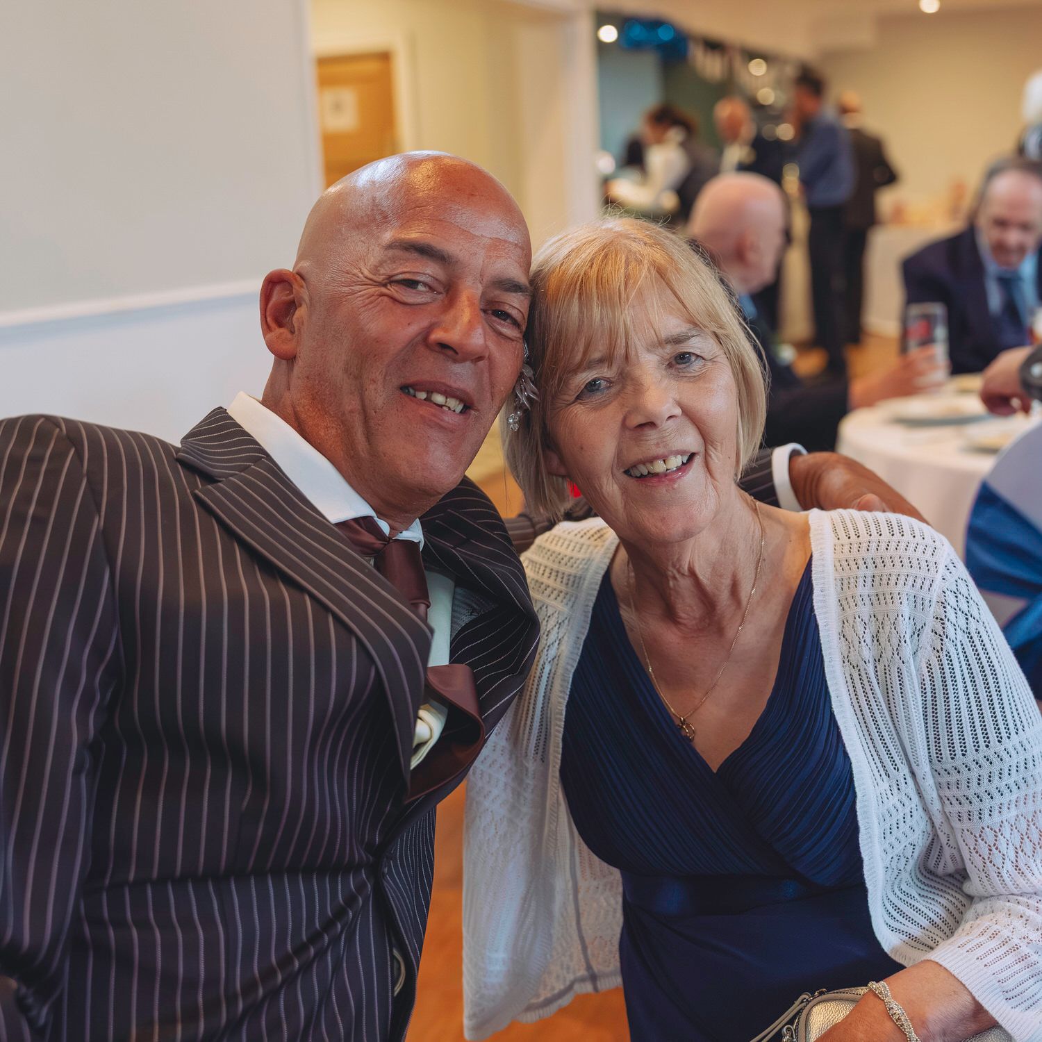 Two smiling adults pose together at a formal event, man in striped suit with tie, woman in navy dress.