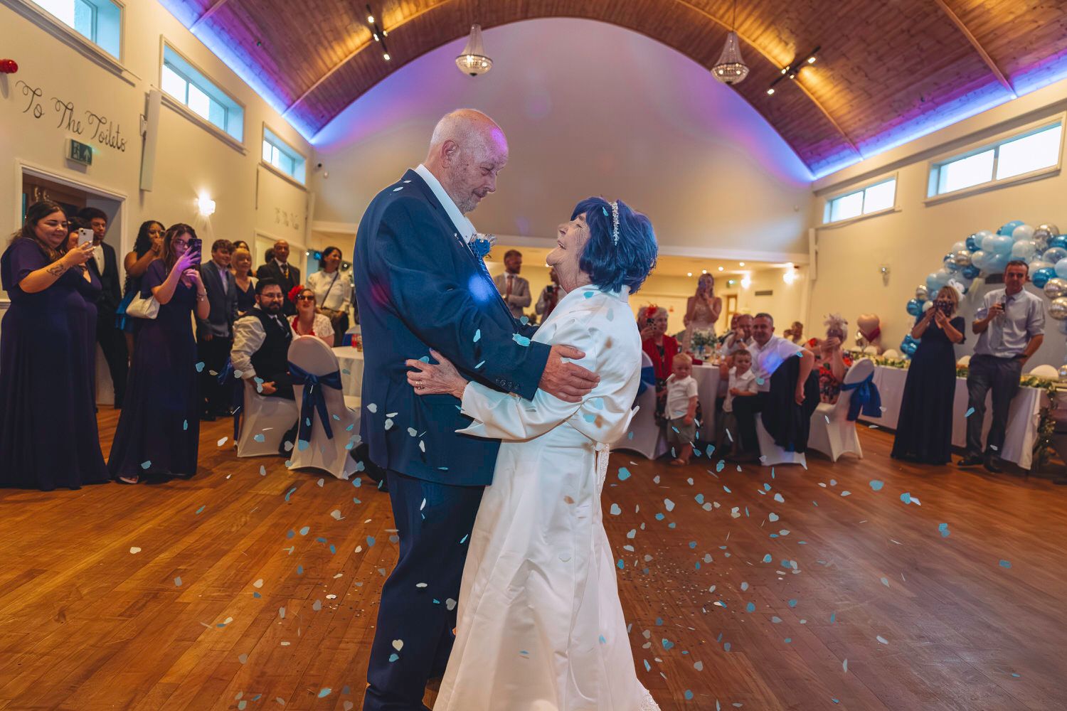Elderly couple sharing their first dance at a wedding reception, surrounded by guests and balloon decorations.