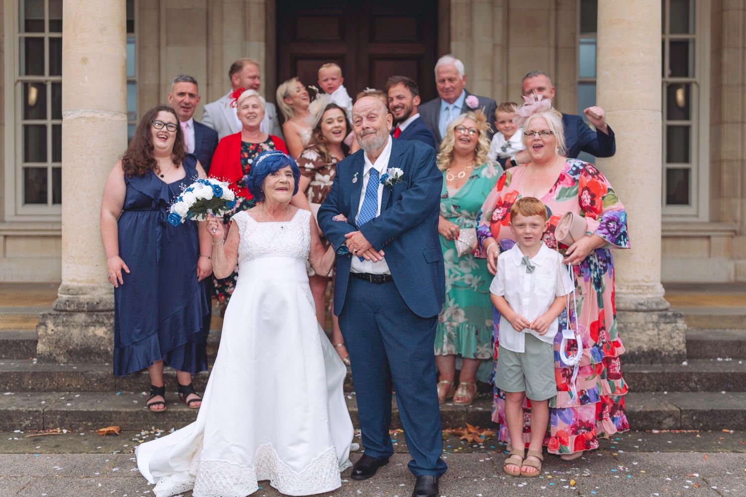 Wedding group photo with bride and groom in white and blue outside a grand building with family and friends.