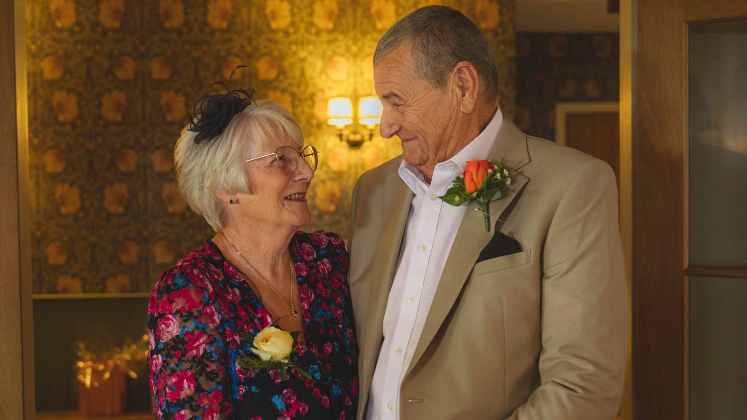 Elderly couple in wedding attire smiling at each other, woman in floral dress, man in beige suit with boutonniere.