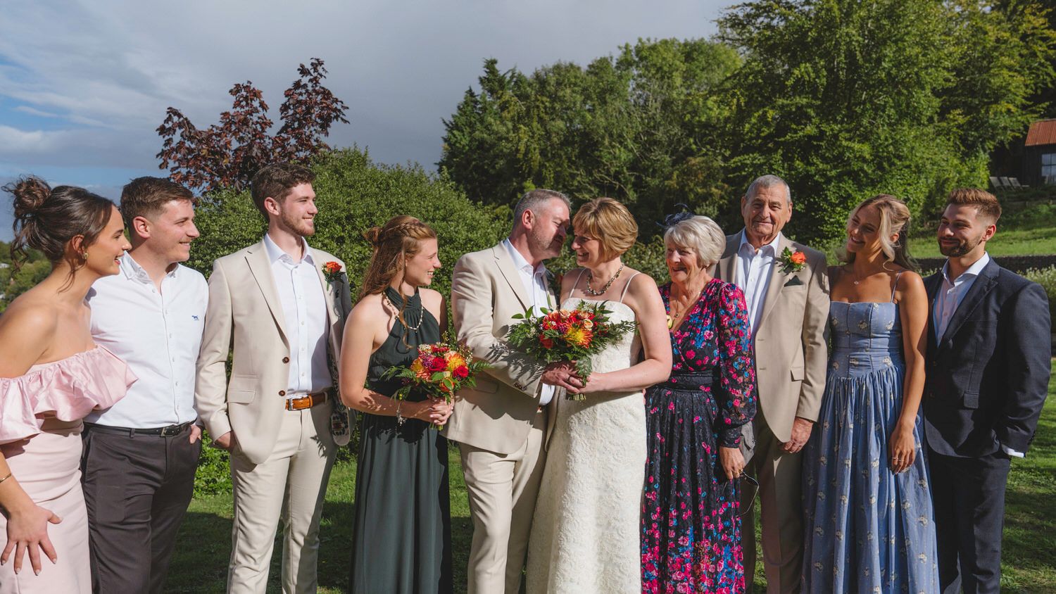 Wedding party group photo outdoors, bride and groom kissing while family celebrates around them in formal attire.